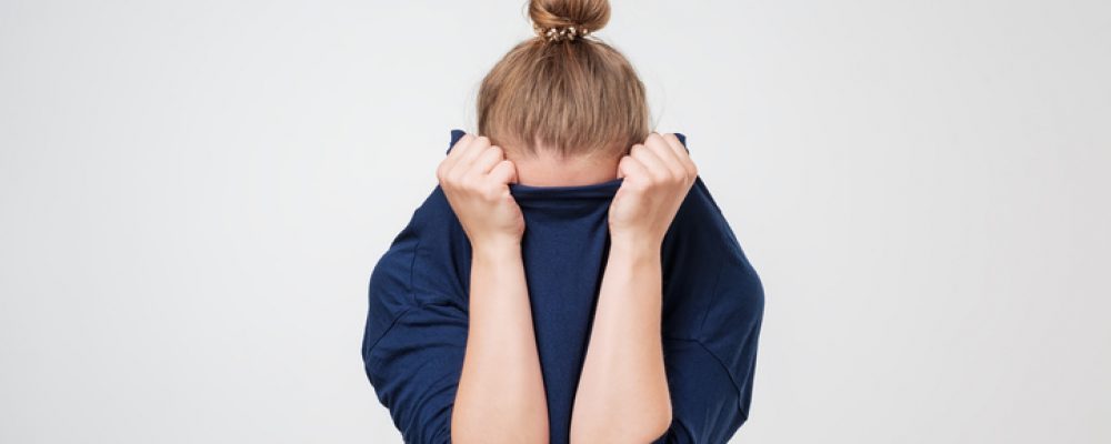 Eine Frau mit Migräne vor grauem Hintergrund, die ihr Gesicht in ihrem dunkelblauen Pullover versteckt.