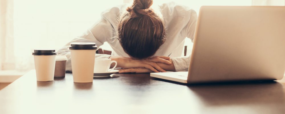 Eine Frau, die mit Migräne an einem Büroschreibtisch sitzt und den Kopf in die Hände gelegt hat.